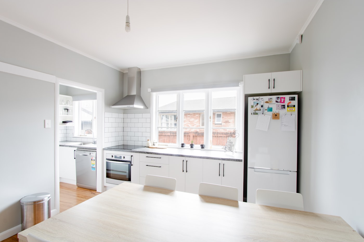 A finished kitchen in an older timber house: white flat-panel cabinetry with black bar handles, subway-tile splashback, and a run of windows over the bench looking onto the neighbouring brick house